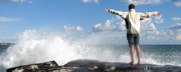 Hombre de espaldas sobre una roca abre los brazos ante las olas, en gesto de liberación