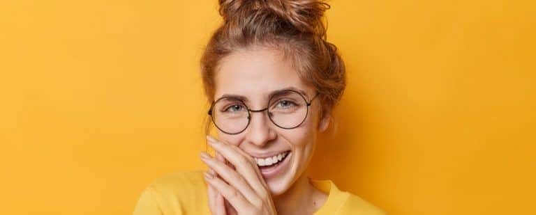 Joven con moño y gafas redondas ríe entusiasmada con las manos junto a la cara