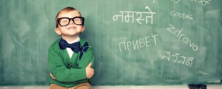 ¿Qué ventajas tiene ser bilingüe? 3 Niño pequeño con gafas y pajarita sonríe delante de una pizarra con saludos en varios alfabetos