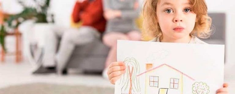 Niña seria muestra su dibujo de una casa mientras dos adultos aparecen sentados y desenfocados detrás