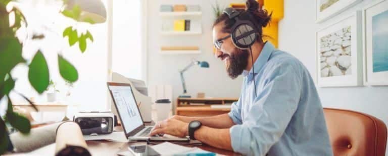 Hombre con auriculares teclea sonriente en un portatil, en un espacio de trabajo montado en casa