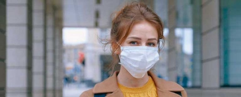 Coronavirus: ¿Consencuencias sobre nuestra salud mental? 1 Mujer joven con mascarilla quirúrgica y abrigo camel mira a cámara en una calle durante la pandemia